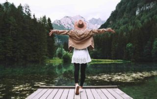 Girls throwing arms out to side on pier and she looks across lake in front of trees and mountains