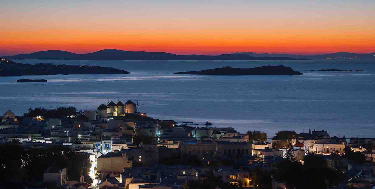 Orange glowing sunset overlooking islands of Mykonos with blue sea and evening lights on windmills and Mykonos buildings