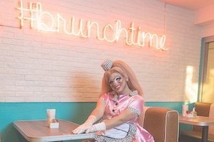 Drag queen smiling at diner restaurant with neon brunch sign illuminated on pink back wall
