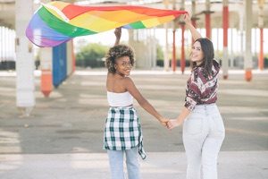 Two girls holding hands and LGBTI flag smiling walking away from camera