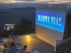 Outdoor cinema screen showing Mamma Mia movie text in restaurant overlooking Santorini Caldera
