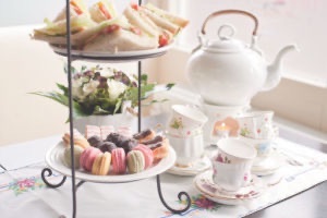 Arrangement of high tea nibbles including macarons, pastries, finger sandwiches & pot of tea and tea cups on side.